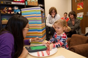 A two-year-old girl looks at a woman giving instructions with researchers watching in the background.