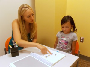 A woman points to explain a workbook game to a child who is watching.