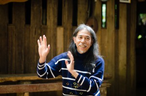 A smiling man in a blue sweater, speaking with gestures.