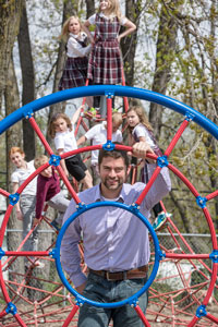 Teacher Zach Steigauf on a playground with children in uniforms on equipment behind him
