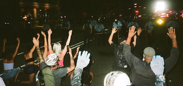 Protestors sit on the street and hold their hands in the air for the riot police