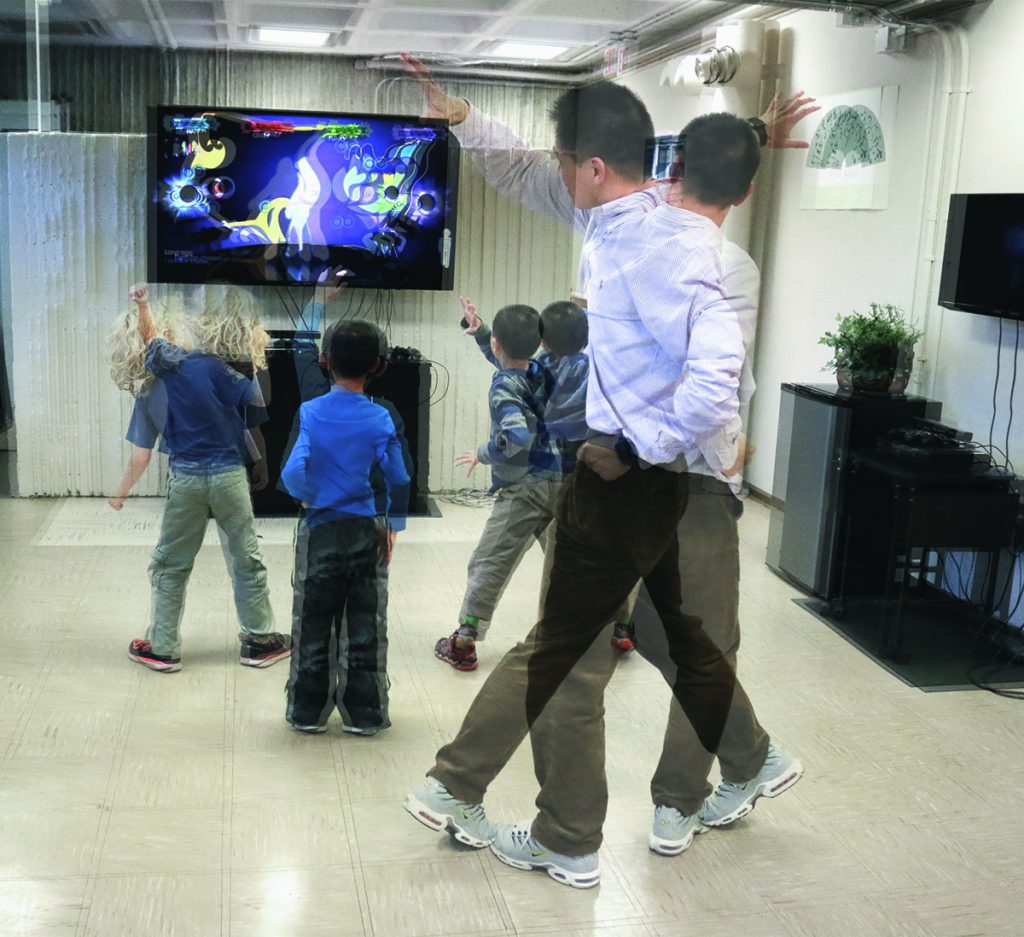 Three children and Zan Gao copy movements on a video screen