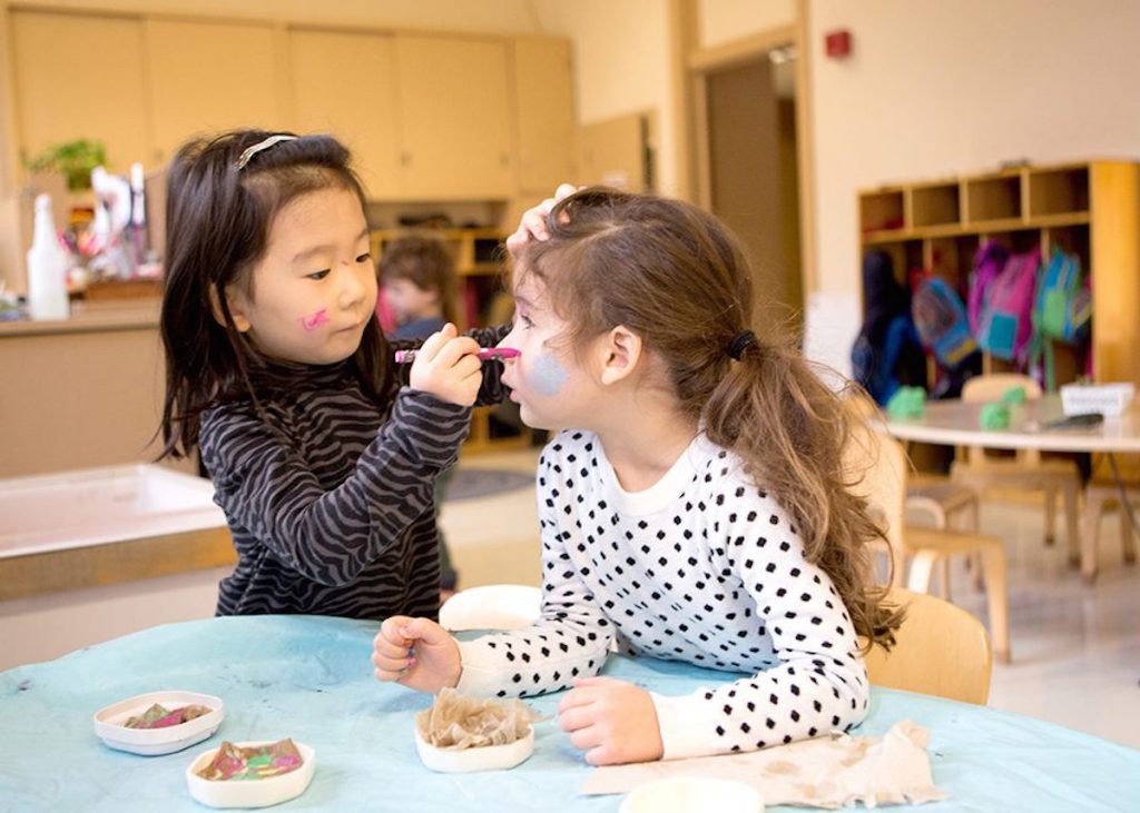 Photo: A preschool girl paints her classmate's face at a classroom table.