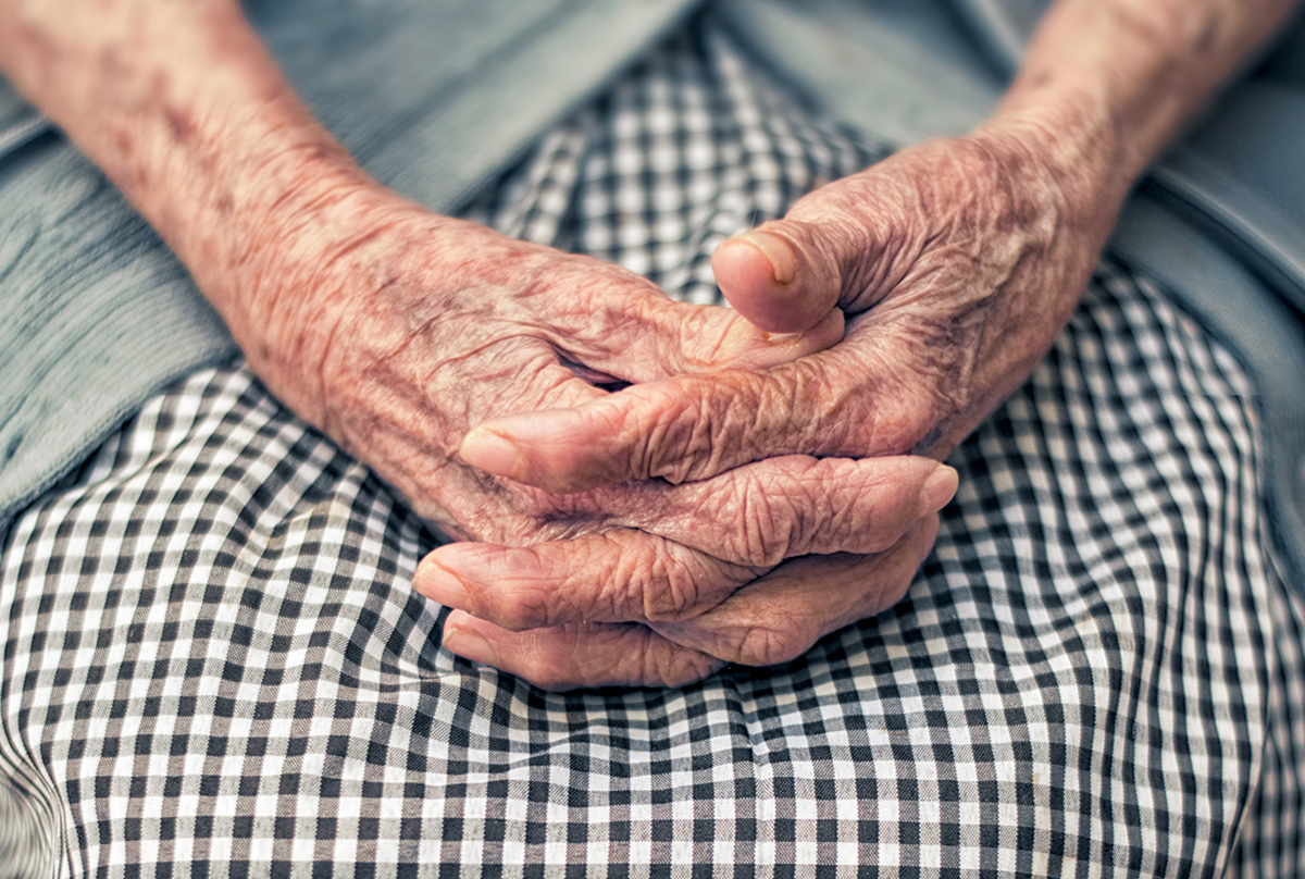An elderly woman folds her hands in her lap.