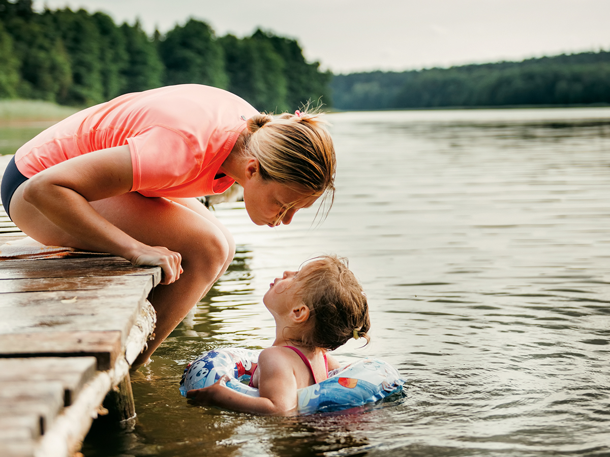 A mother leans over a pier as her daughter swims with an innertube in a lake.