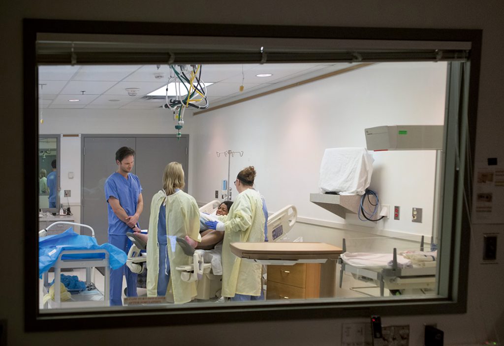 Viewed through a window, three students participate in a medical simulation with a standardized patient portraying a woman in labor