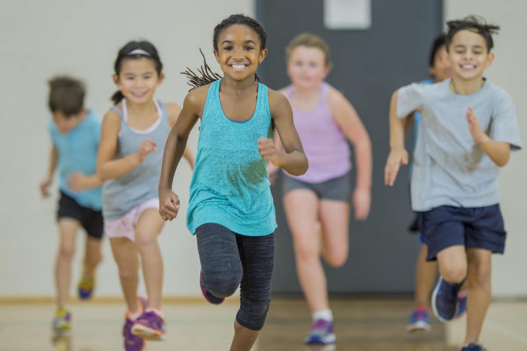 A multi-ethnic group of elementary school students are indoors in a school gym. They are wearing athletic clothing and shoes. They are jogging towards the camera.