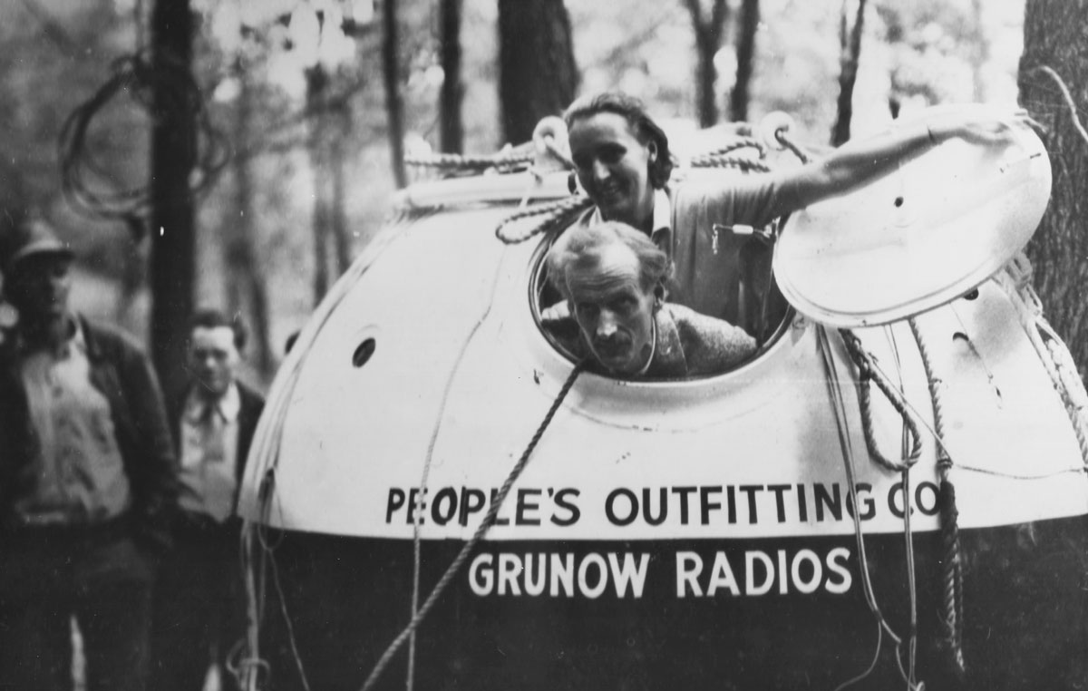 Jean and Jeannette Piccard opening the hatch of a pressurized 7-foot sphere in a wooded landscape